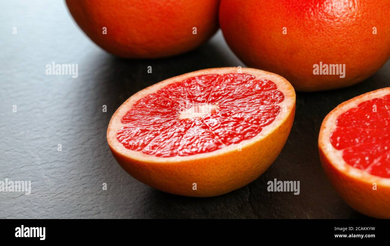 Nahaufnahme - Hälfte der rosa Grapefruit auf dunklem Arbeitsbrett, mehr Zitrusfrüchte im Hintergrund Stockfoto