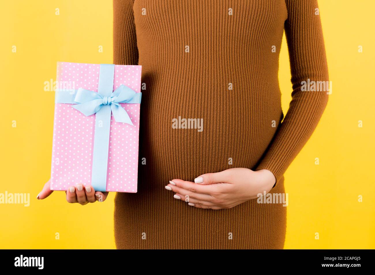 Zugeschnittenes Bild von rosa gefleckte Geschenkbox in der Hand der schwangeren Frau gegen ihren Bauch auf gelbem Hintergrund. Zukünftige Mutter in braunem Kleid. Warten auf einen BA Stockfoto