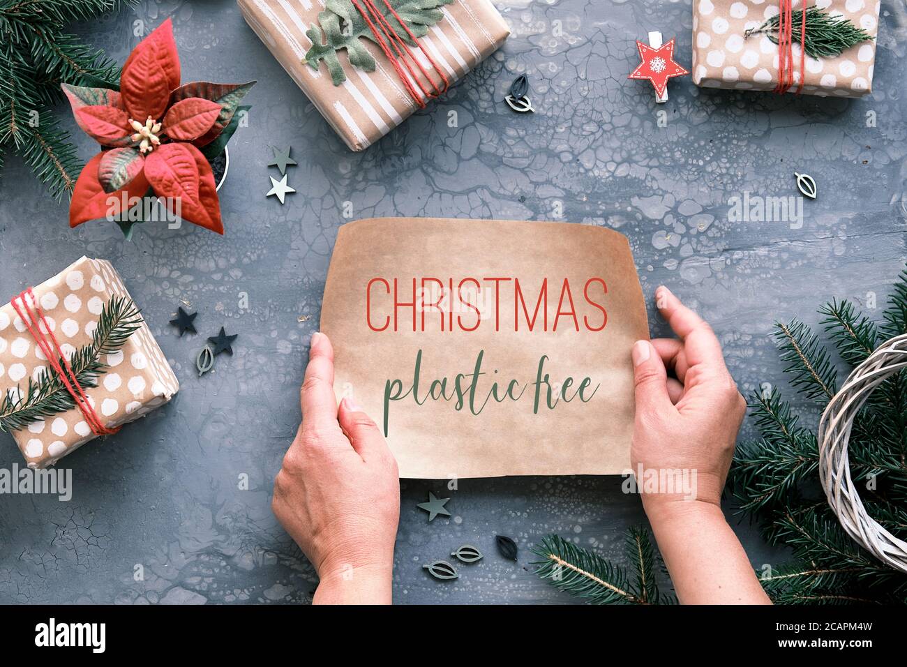 Text Weihnachten Kunststoff frei auf Stück Handwerk Geschenkpapier in den Händen. Flach legen auf grauen Tisch mit Weihnachtsgeschenke in handgefertigtem braunem Papier verpackt Stockfoto
