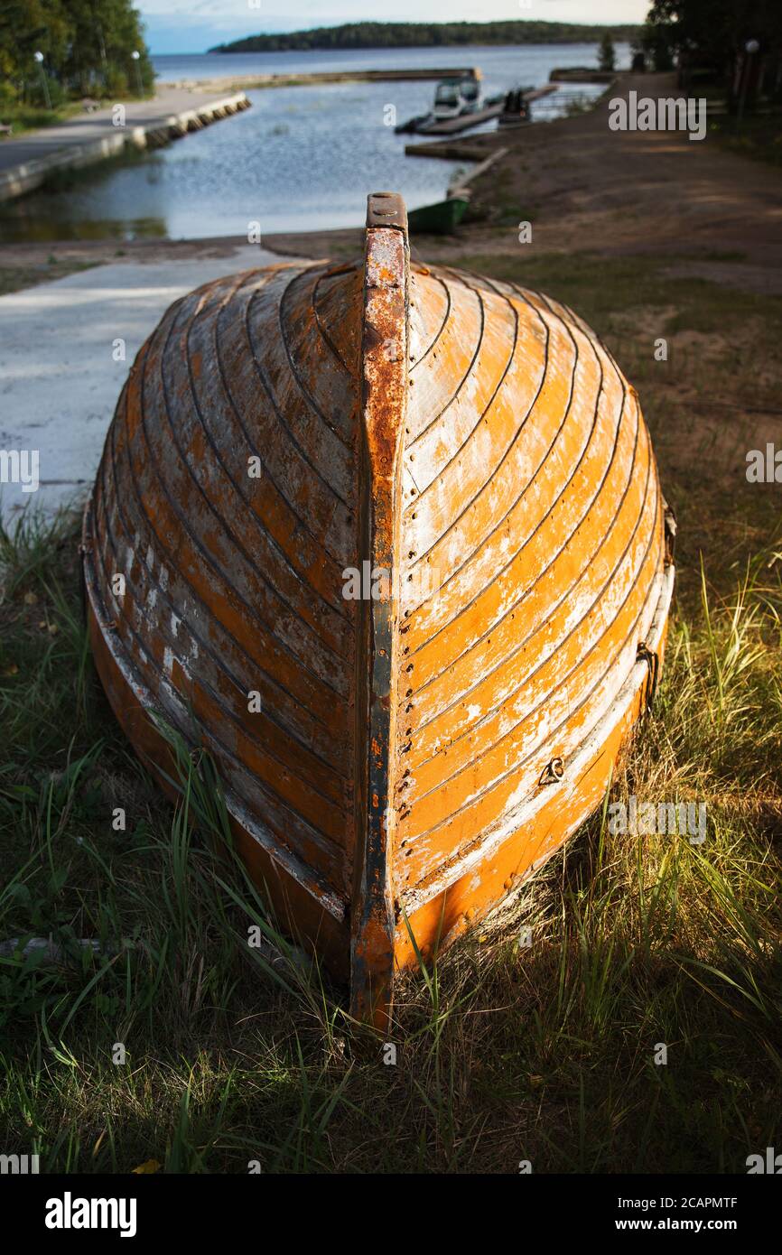 Altes schäbiges orangefarbenes Holzboot zur Restaurierung, das von der Sonne auf altem trockenen Gras beleuchtet wird, rissige Farbe auf dem Körper. Pier, ruhige Bucht und wolkige Herbst s Stockfoto