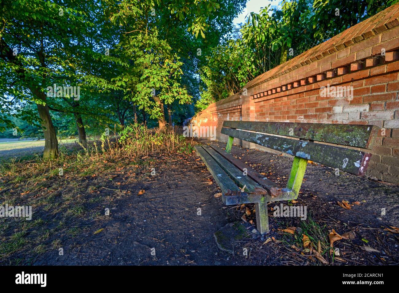 Eine alte und kaputte Bank in Hardington Park, Beckenham, Kent. Die Bank ist bei Sonnenuntergang mit einer roten Ziegelmauer und Bäumen dahinter. Stockfoto