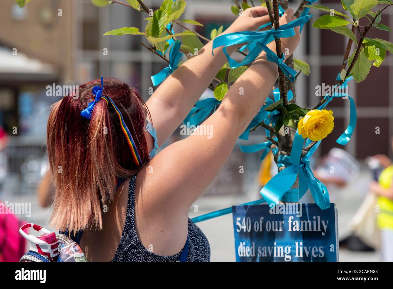 Eine Frau mit Regenbogenhaarbändern bindet ein blaues Band an einen Baum, um das von COVID-19 in Basildon, Essex, Großbritannien, getötete Gesundheitspersonal zu ehren. Geblümter Tribut Stockfoto