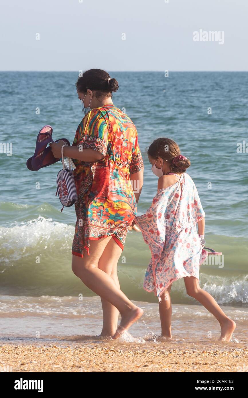 Punta Umbria, Huelva, Spanien - 7. August 2020: Mutter und Tochter gehen am Strand mit schützenden oder medizinischen Gesichtsmasken spazieren. Neues normales Spa Stockfoto