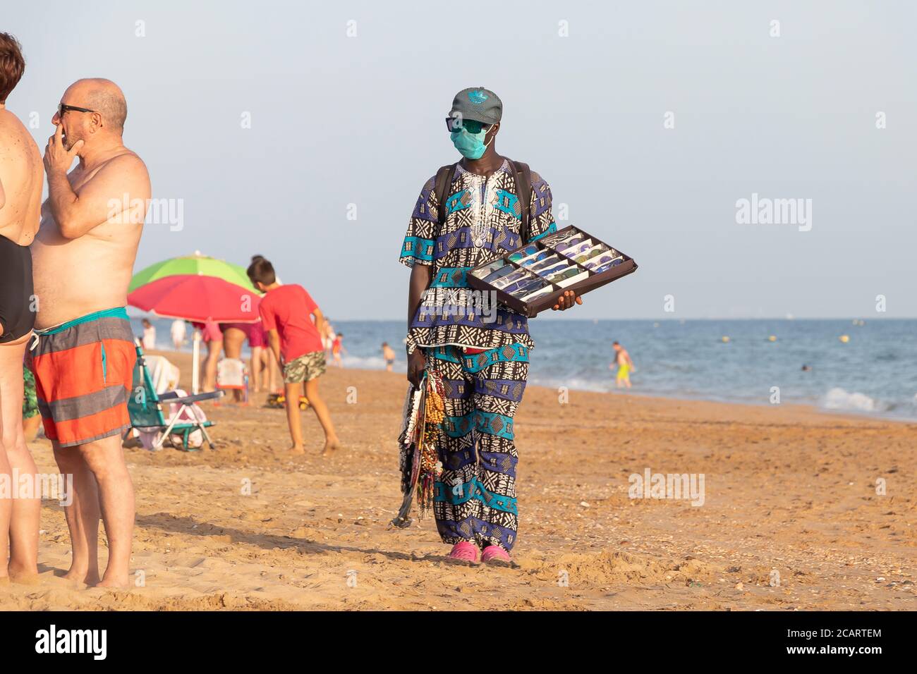 Punta Umbria, Huelva, Spanien - 7. August 2020: Afrikanische Einwanderer verkaufen ihre Waren am Strand mit schützenden oder medizinischen Gesichtsmaske. Alle Waren von Stockfoto