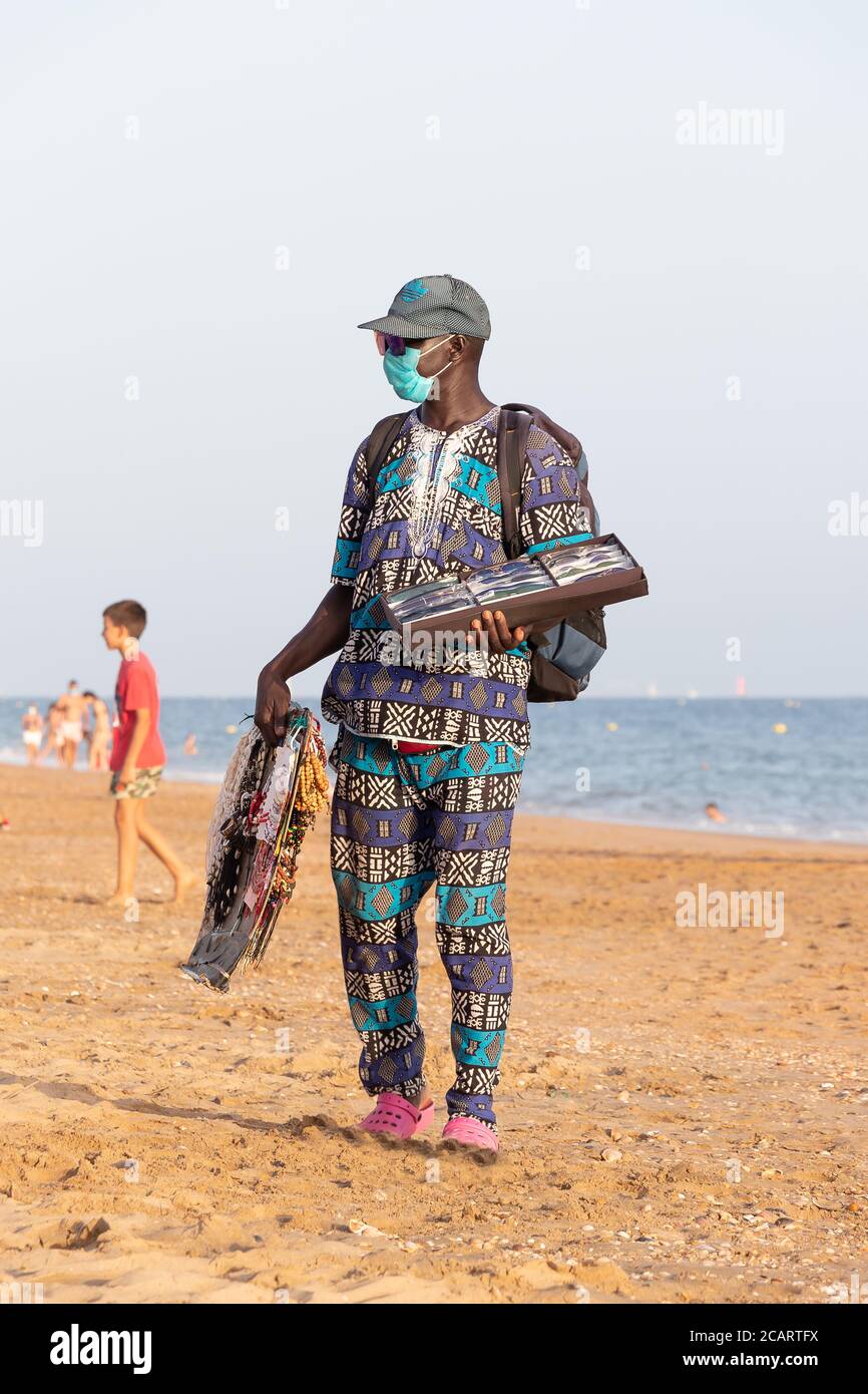 Punta Umbria, Huelva, Spanien - 7. August 2020: Afrikanische Einwanderer verkaufen ihre Waren am Strand mit schützenden oder medizinischen Gesichtsmaske. Alle Waren von Stockfoto