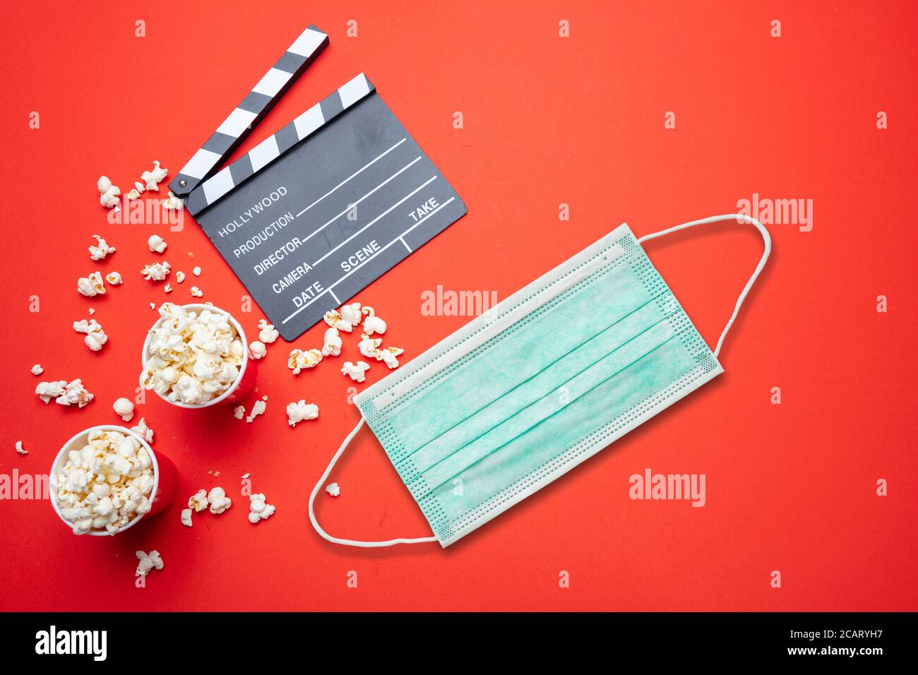 Coronavirus-Schutz im Kino. Medizinische Schutz Gesichtsmaske, Film Clapperboard und Pop Mais vor rotem Hintergrund. COVID-19-Krankheit Stockfoto