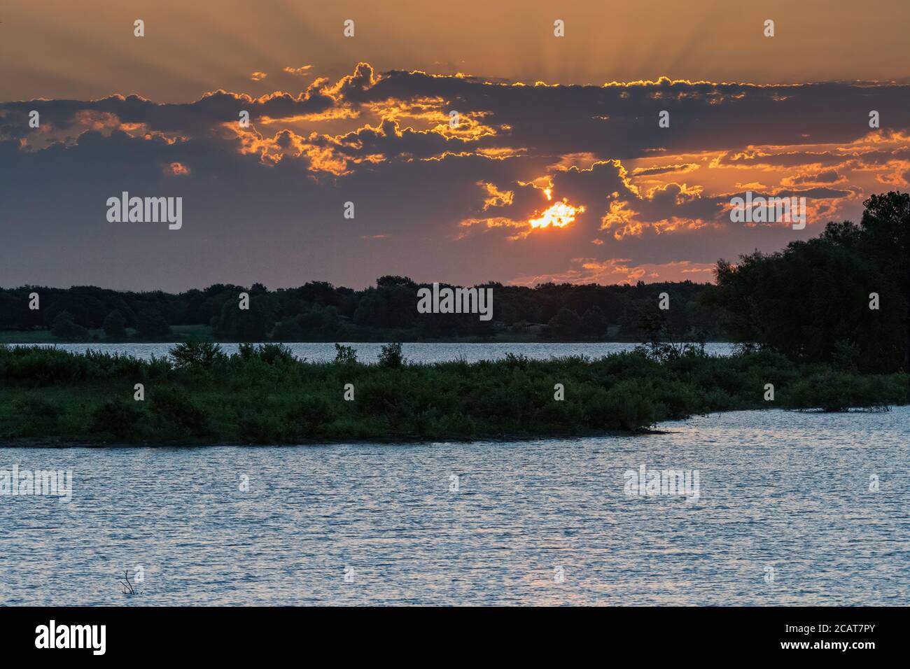 Sonne späht durch die frühen Morgenwolken über einem See mit einer von Bäumen bedeckten Halbinsel, die das Wasser in zwei Seen teilt. Stockfoto