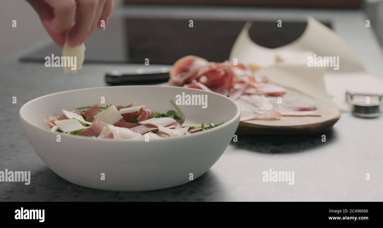 Man Hand hinzufügen Parmesan Flocken Salat mit Rucola, Tomaten und Prosciutto zu machen Stockfoto