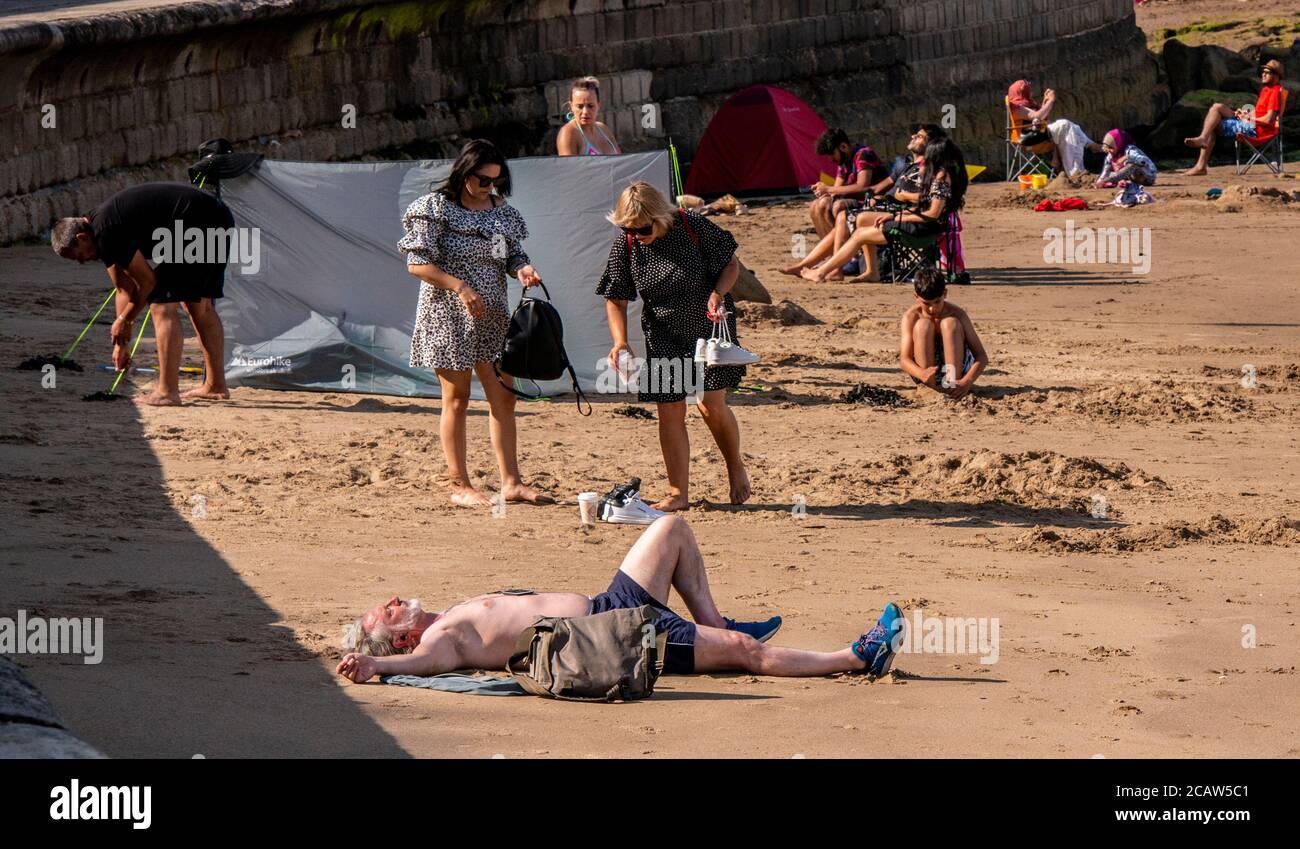 Ein Mann schläft am Scarborough Beach mit ausgestreckten Armen und anderen Strandbesuchern im Hintergrund. Stockfoto
