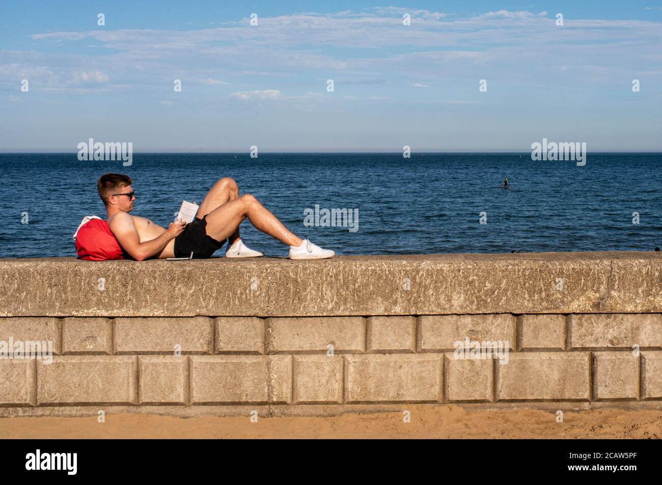 Ein junger Mann legt sich beim Lesen auf eine niedrige Wand Ein Buch mit dem blauen Meer im Hintergrund in Scarborough Stockfoto