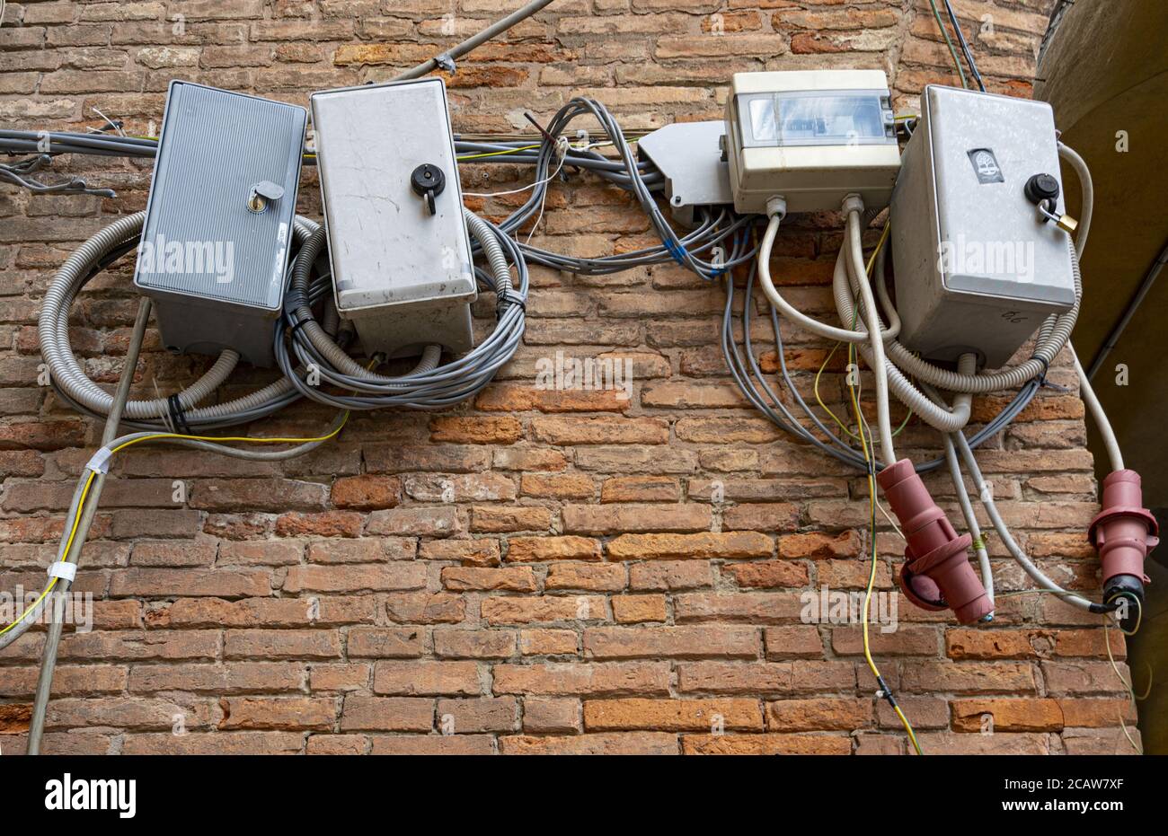 Ferrara, Italien. 6. August 2020. Chaotische Verbindungen und Verteilerkästen von elektrischen Kabeln an einer Ziegelwand Stockfoto