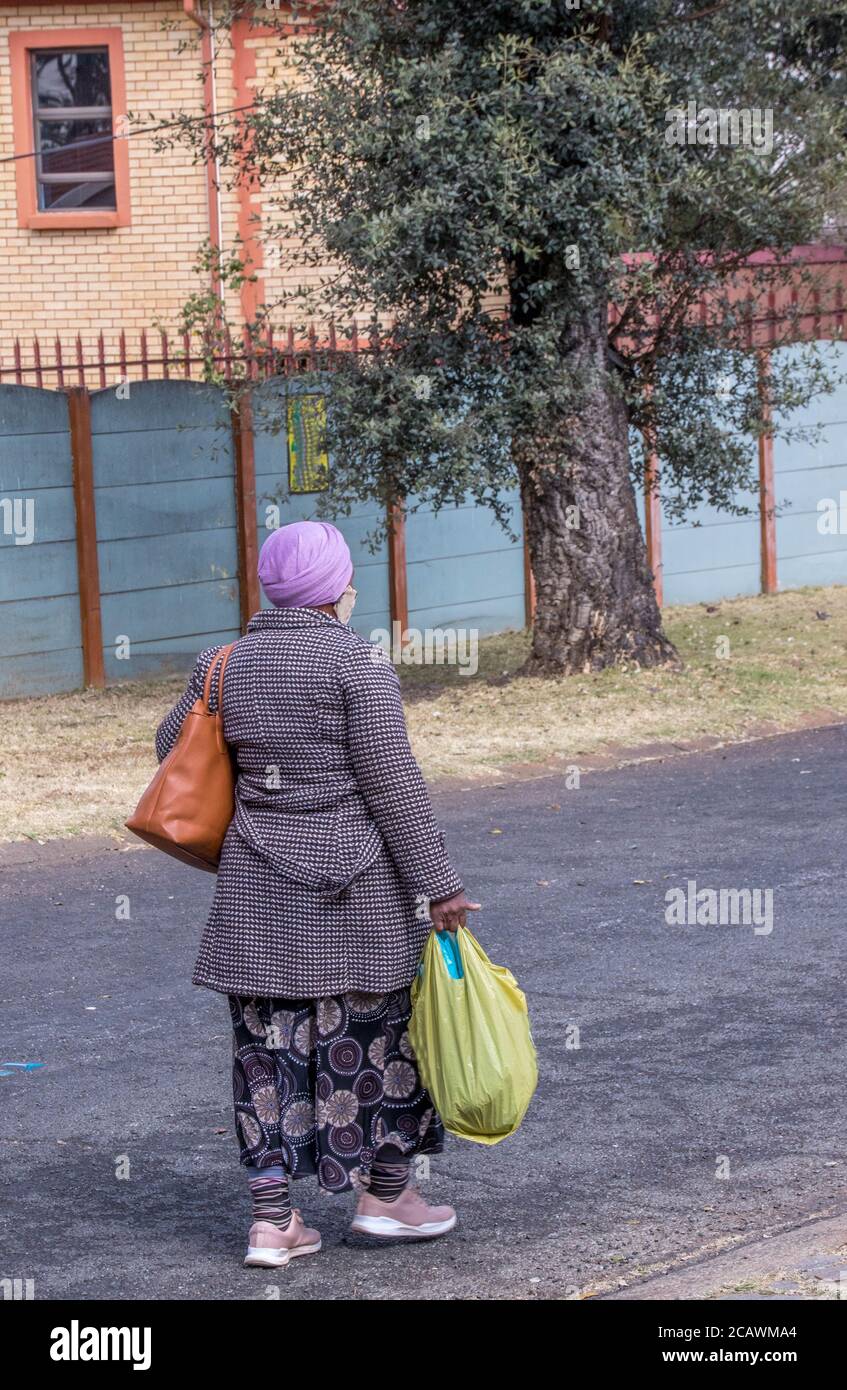 Alberton, Südafrika - eine schwarze Frau mit Gesichtsmaske geht mit ihren für kaltes Wetter gekleideten Einkäufen vom Laden nach Hause Stockfoto