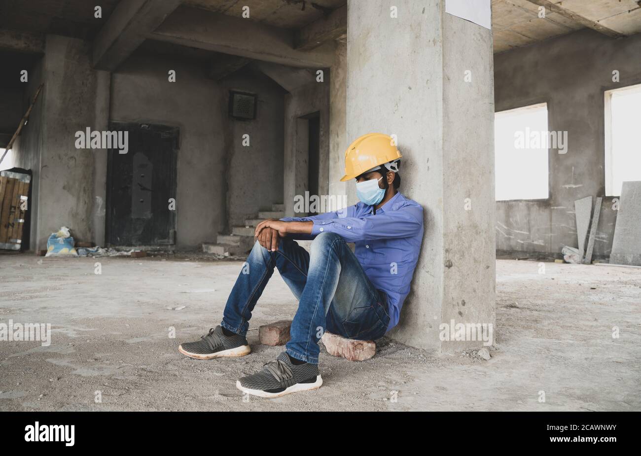 Bauarbeiter sitzt traurig und einsam auf der Baustelle trägt eine medizinische Maske mit Hardhut, um zu verhindern, Covid-19 Verbreitung, Konzept der Arbeitslosigkeit Stockfoto