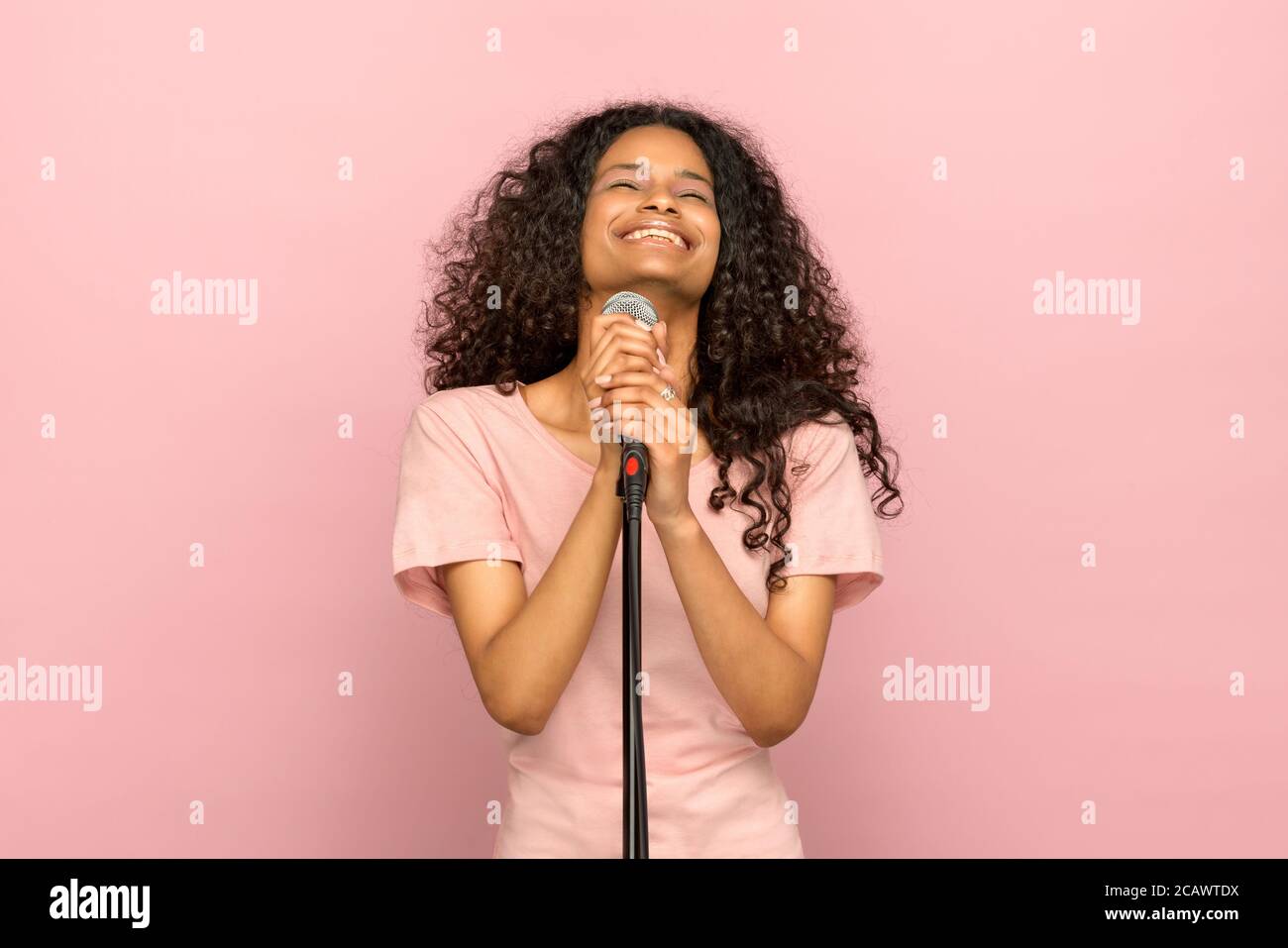 Lächelnd glücklich junge schwarze Mädchen singen in ein Mikrofon mit Kopf nach hinten gekippt und strahlendes Lächeln gegen ein rosa Studio Hintergrund Stockfoto