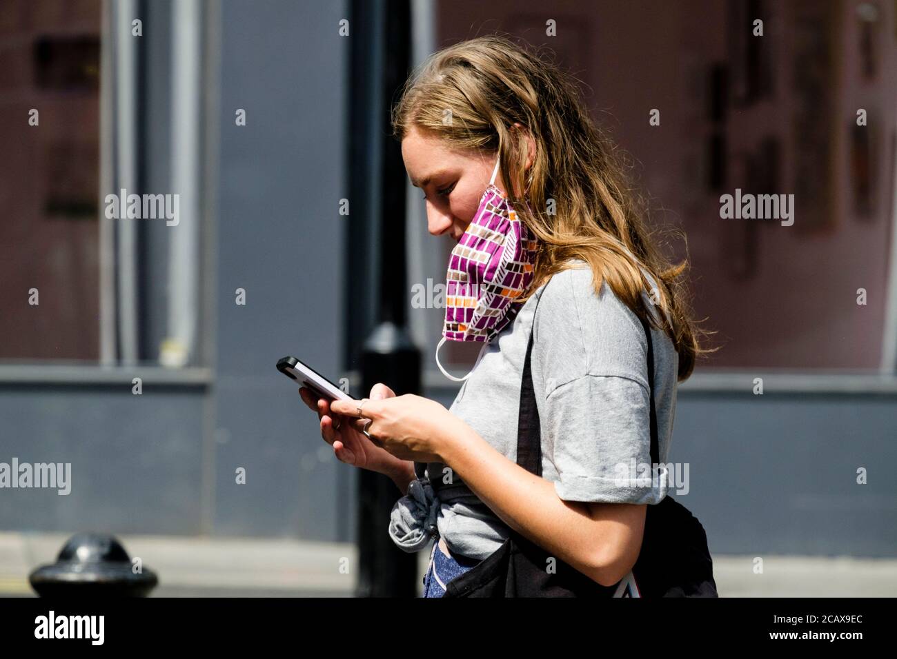 Die junge Frau mit Gesichtsbedeckung, die an einem Ohr hängt, nutzt das Smartphone auf der Londoner Straße. Stockfoto
