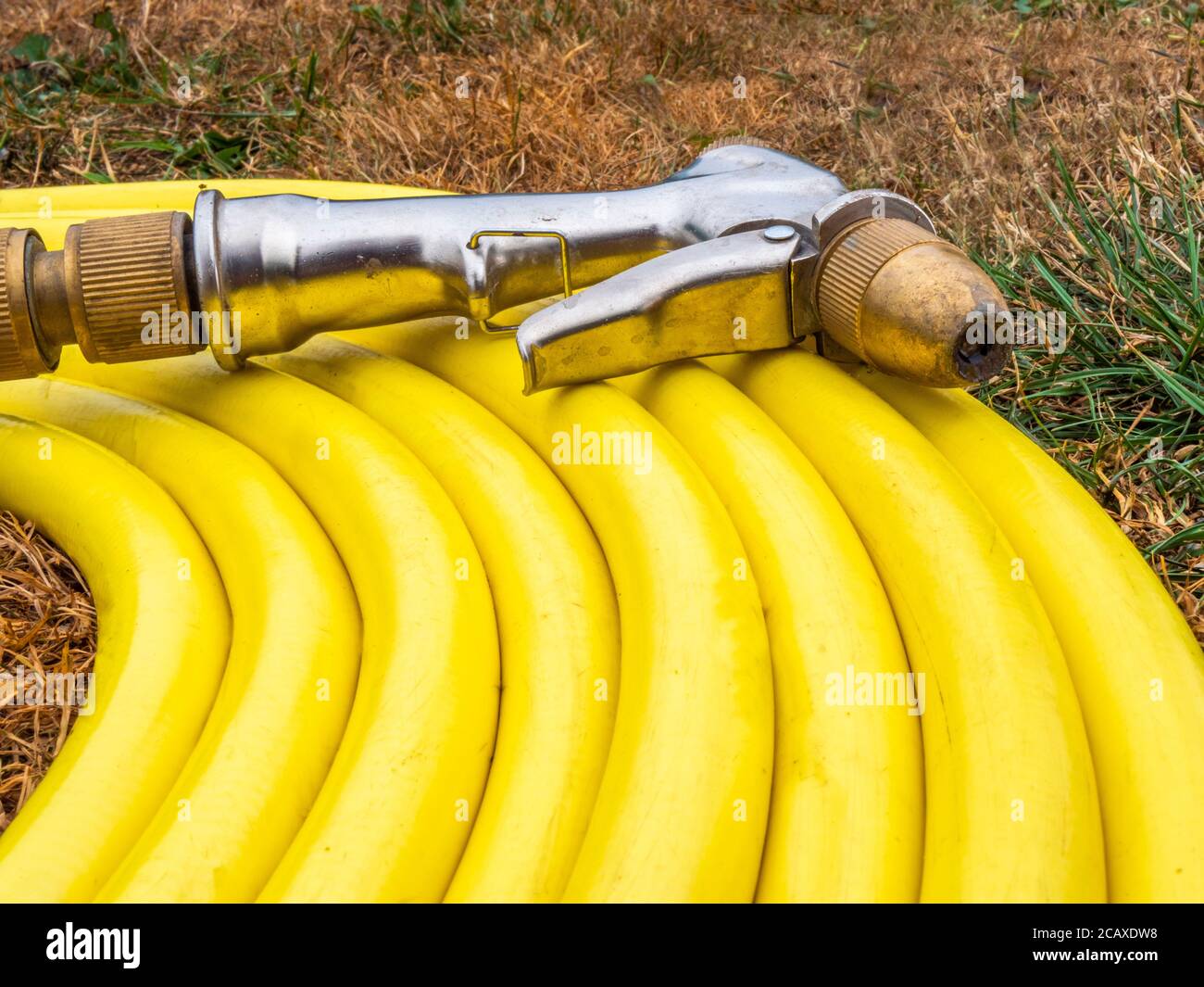 Ein gelber Wasserschlauch ohne Brandmarke, mit angefügter Spritzpistole, auf ausgetrockentem braunem Gras in einem Gebiet, in dem ein Wassermangel zu einem Schlauchleitungsverbot geführt hat. Stockfoto