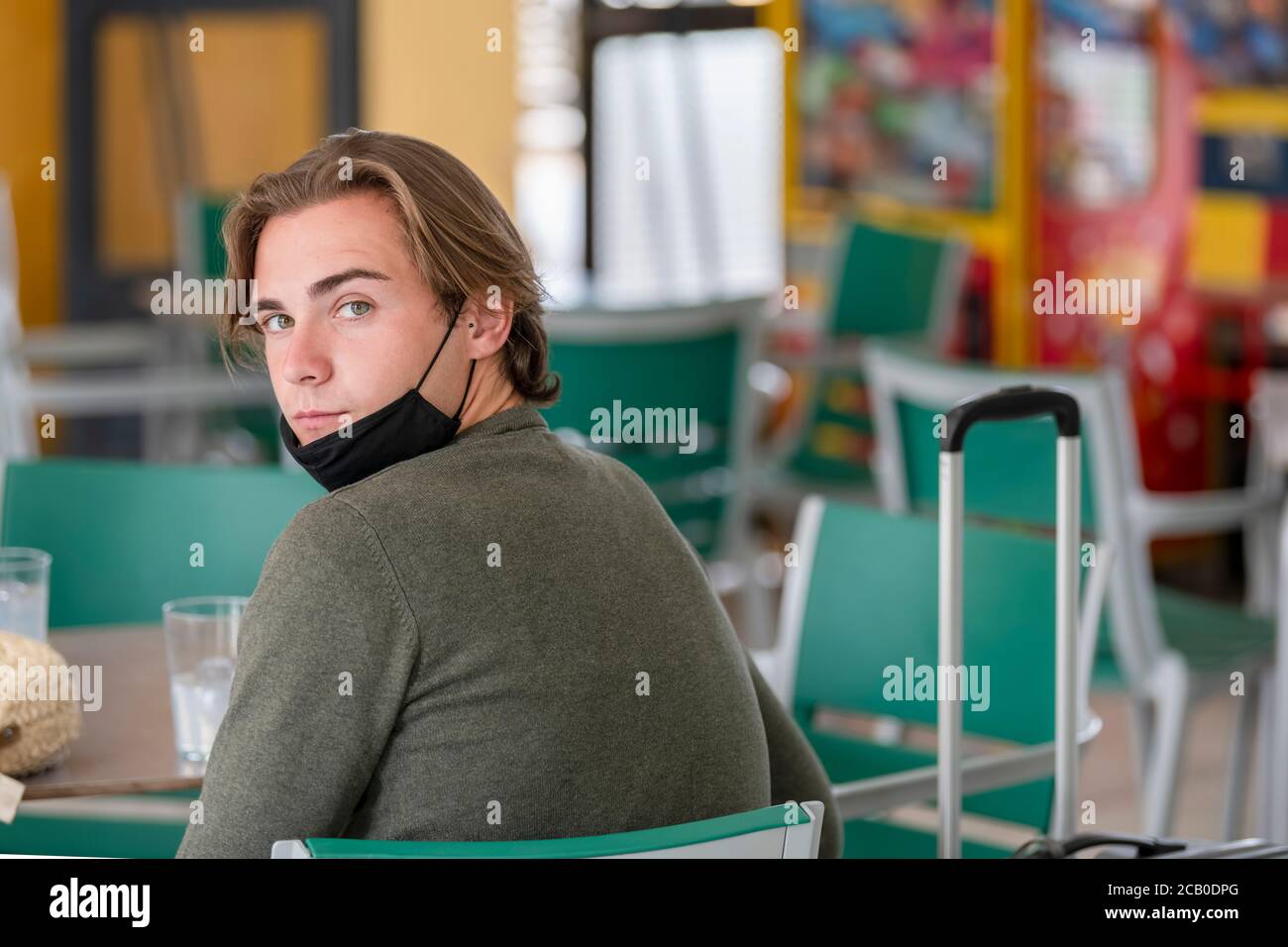 Nahaufnahme eines hübschen jungen Mannes mit heruntergezogener Gesichtsmaske und Blick zurück auf die Kamera, während er in einer Cafeteria am Bahnhof wartet. Reisen und sicher Stockfoto