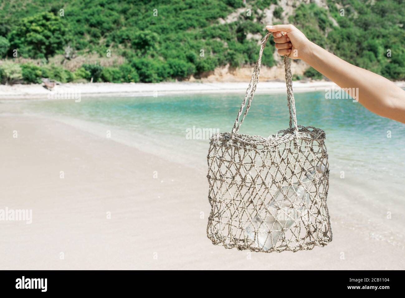 Frau hält Stroh Weidenwiederverwendbare Tasche mit Glasflasche für Wasser. Plastik frei und Zero Waste Lifestyle. Weißer Sandstrand mit klarem türkisfarbenem Wasser im Hintergrund. Stockfoto