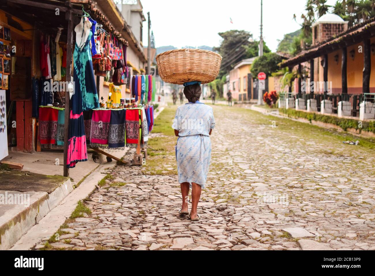 Concepcion de Ataco / El Salvador - 29. Oktober 2017: Salvadorianische Frau, die auf einem Steinweg mit einem Korb auf dem Kopf geht und Soldaten im Hintergrund patrouillieren Stockfoto