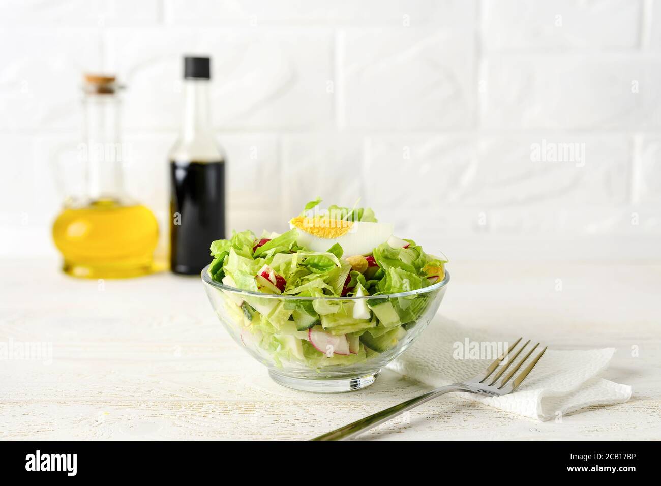Frisch hausgemachter Salat aus Salat, Gurke, Rettich und gekochten Eiern in einer Glasschüssel auf einem weißen Holztisch. Olivenöl und Sojasauce zum Dressing. Stockfoto