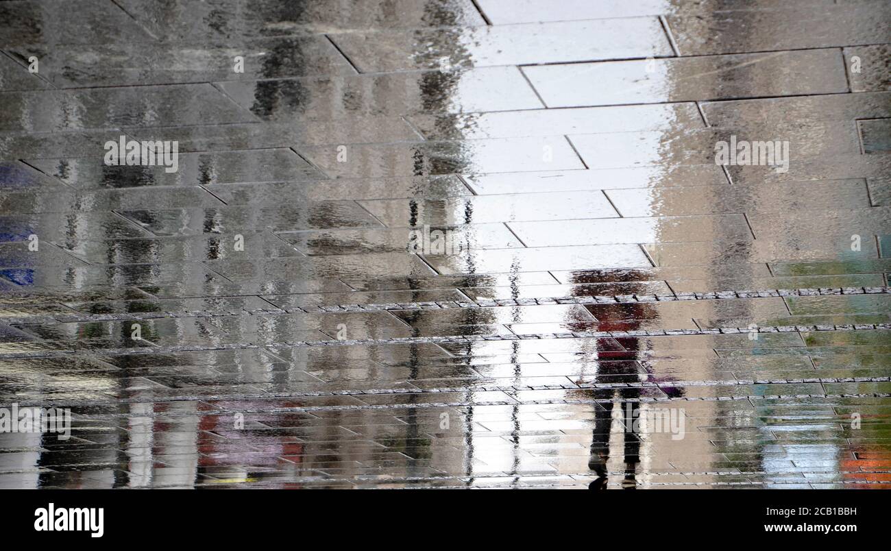 Verschwommener Spiegelschatten Silhouette auf nasser Pfütze einer Frau, die mit Jacke über ihr auf einer Stadtstraße an einem regnerischen Tag ging, abstrakter Hintergrund Stockfoto