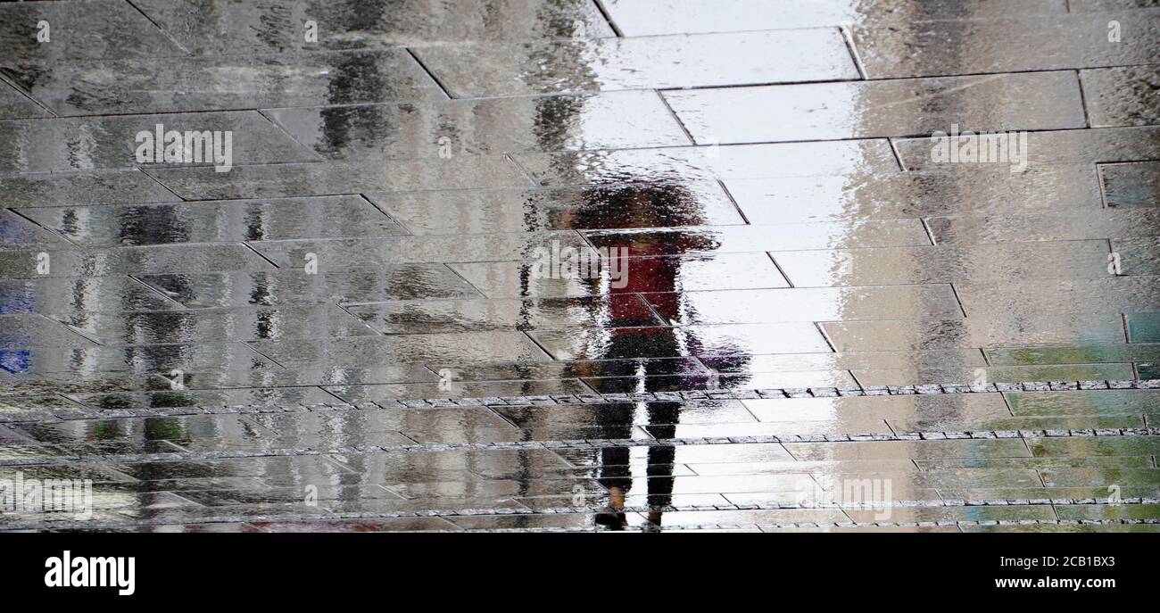 Verschwommener Spiegelschatten Silhouette auf nasser Pfütze einer Frau, die mit Jacke über ihr auf einer Stadtstraße an einem regnerischen Tag ging, abstrakter Hintergrund Stockfoto