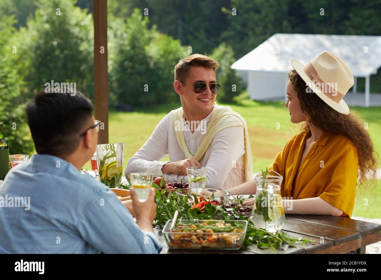 Buntes Porträt eines lächelnden jungen Paares, das miteinander spricht, während man das Abendessen mit Freunden im Freien während der Sommerparty genießt, Platz kopieren Stockfoto