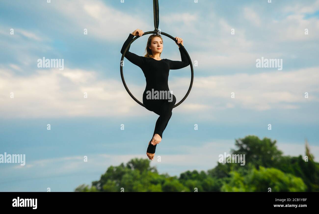 Frau Aerialistin führt akrobatische Elemente in hängenden Luftreifen vor dem Hintergrund des blauen Himmels, weiße Wolken und Bäume. Stockfoto