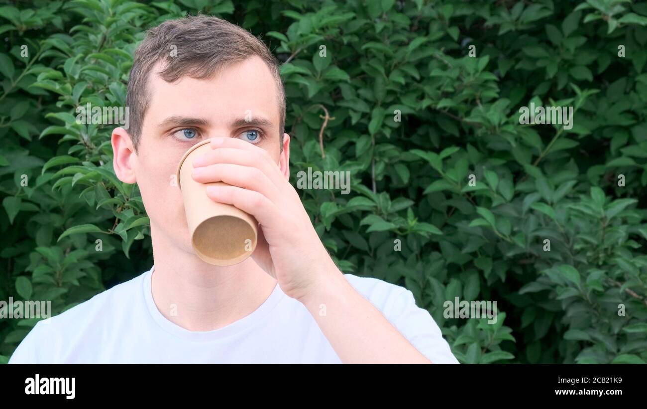Mann mit blauen Augen trinkt Kaffee aus einem Pappbecher Auf dem Hintergrund der Büsche Stockfoto