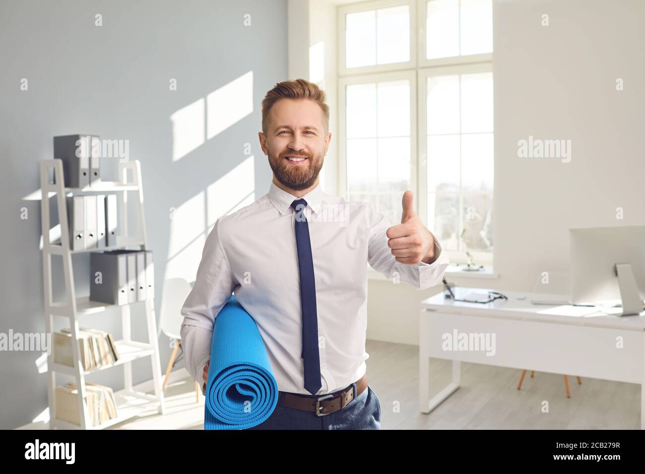 Glücklicher erfolgreicher Geschäftsmann in einem weißen Hemd mit Yogamatte im Büro. Stockfoto