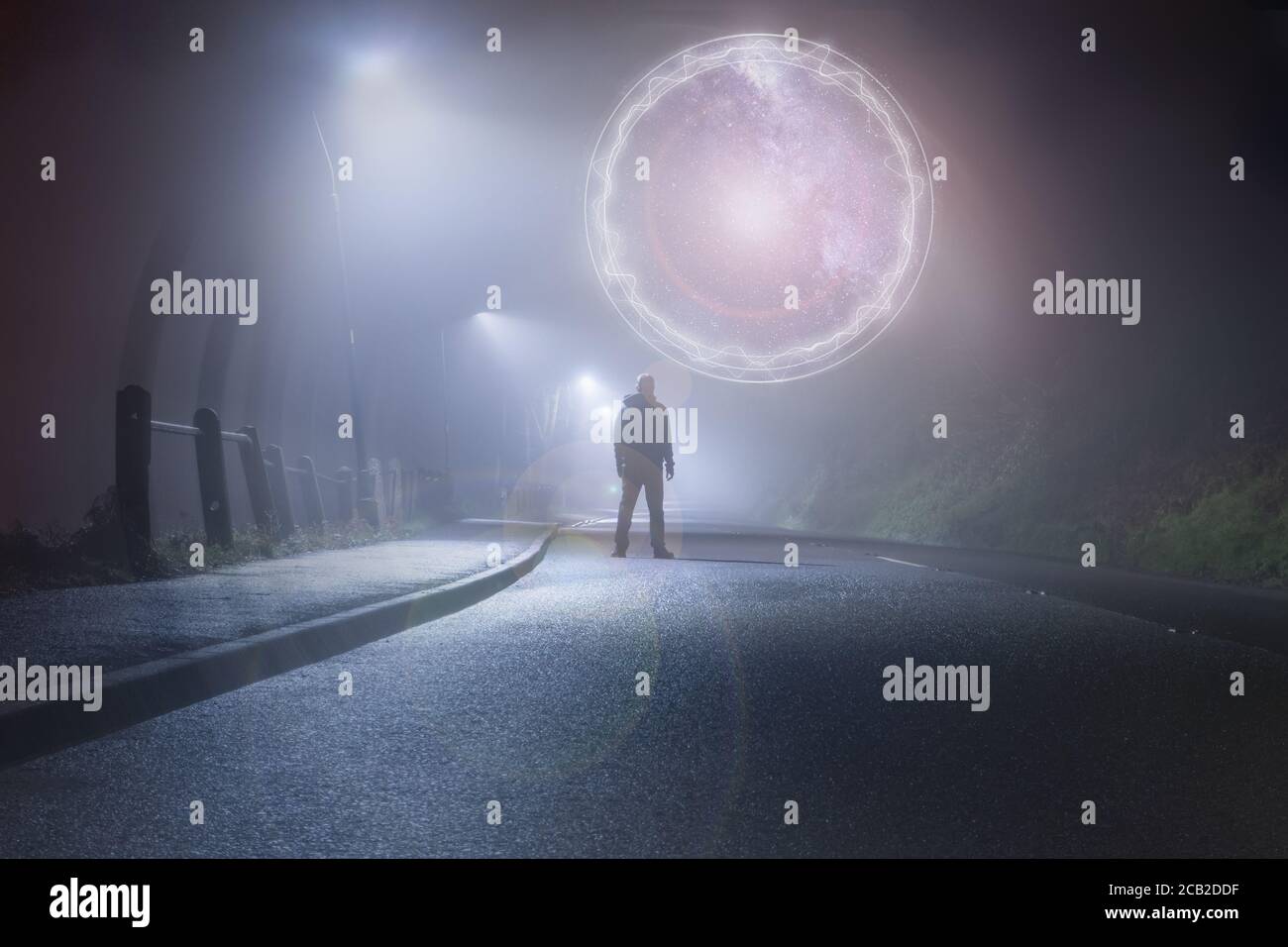Ein Mann, der mitten auf einer Landstraße steht. Unter der Straßenbeleuchtung. In einer nebligen, launischen, gruseligen Winternacht. Blick auf ein glühendes Portal. Stockfoto