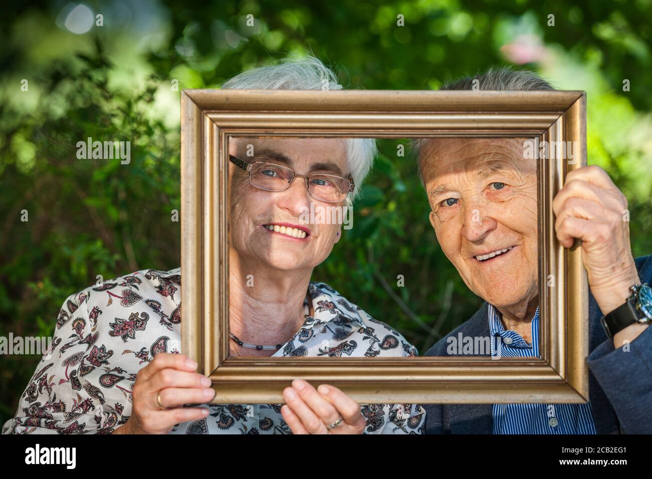 Frontalansicht eines lachenden Rentnerpaares, das durch einen Bilderrahmen vor einer verschwommenen Hecke schaut. Stockfoto