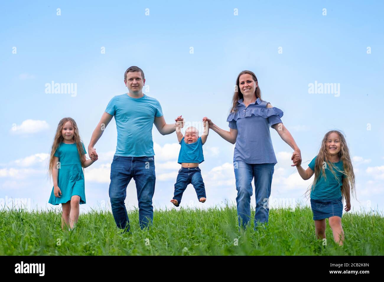 Große Familie Vater, Mutter und drei Kinder gehen auf grünem Gras gegen blauen Himmel. Glückliche kaukasische Eltern, zwei Töchter und ein junger Sohn halten die Hände Stockfoto