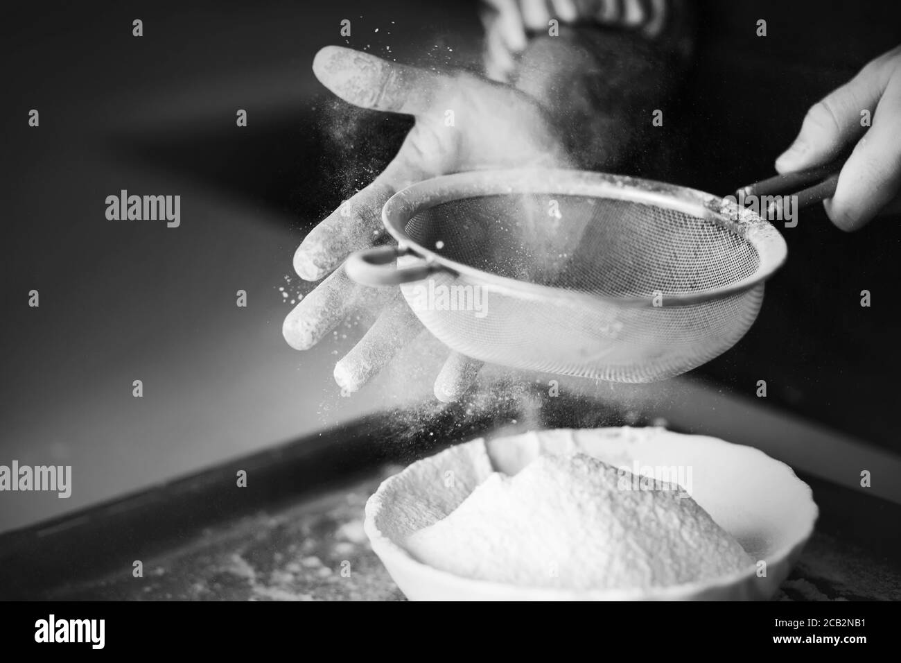 Ein schwarz-weißes Bild eines erfahrenen Küchenchefs, der mit seiner Hand Mehl durch ein Sieb in eine Schüssel sieben lässt. Der Prozess des Kochens. Stockfoto