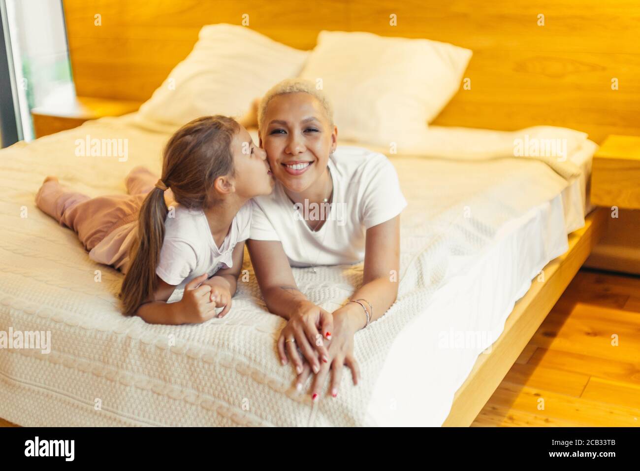 Nette Tochter küssen ihre Mama auf dem Bett liegend. Porträt von glücklichen Eltern und Kind. Liebe, Familienkonzept Stockfoto