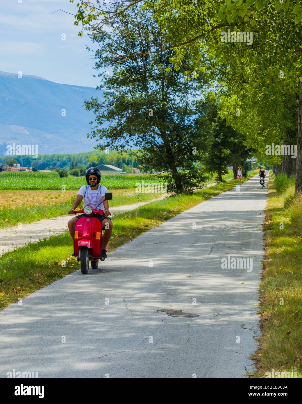 Ein Mann auf einem roten Roller fährt auf einer kleinen Straße durch die Aue des Flusses Topino, in der Nähe von Bevagna, Umbrien, Italien Land Stockfoto