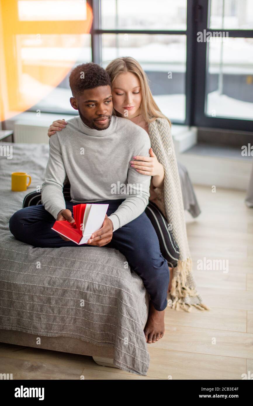 Attraktives gemischtes Paar sitzt zusammen auf dem Bett und liest zusammen ein Buch. Kaukasische blonde Frau trägt warme Wrap auf den Schultern, umarmt ihr afrika Stockfoto