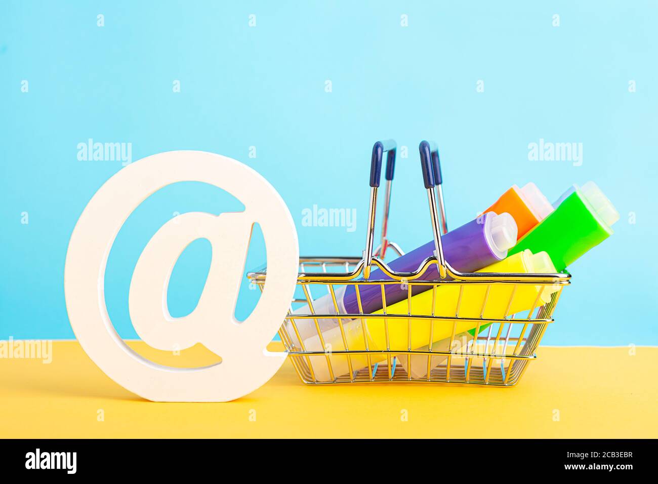 Ein Warenkorb mit Textmarker Stift und AT-Symbol oder Werbespot auf gelbem und blauem Hintergrund. Zurück zur Schule Stockfoto