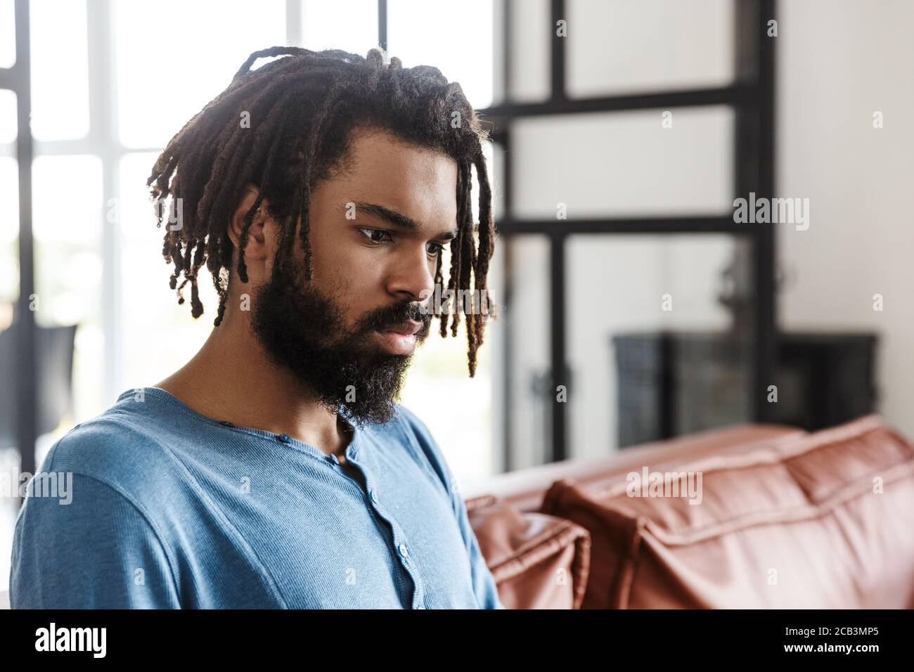 Hübscher junger afrikanischer Mann, der zu Hause auf einer Ledercouch sitzt und wegschaut Stockfoto