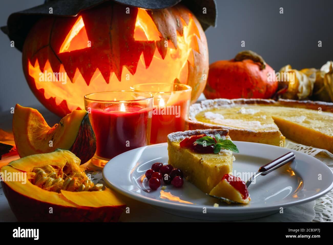 Stück süße hausgemachte halloween Kürbiskuchen mit Minzeblatt und Preiselbeermarmelade auf weißem Teller Nahaufnahme. Kerzen und Jack-o-Laterne im Hut auf dem Hintergrund. Traditionelles Urlaubsessen Stockfoto