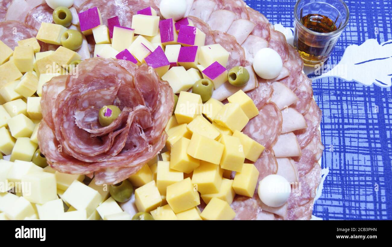 Weißer Teller, verziert mit Salami, Schinken, Wachteleiern und Palmenherzen, auf einem Hintergrund mit blauem Hintergrund, mit einem Whiskey-Glashalter, Stockfoto