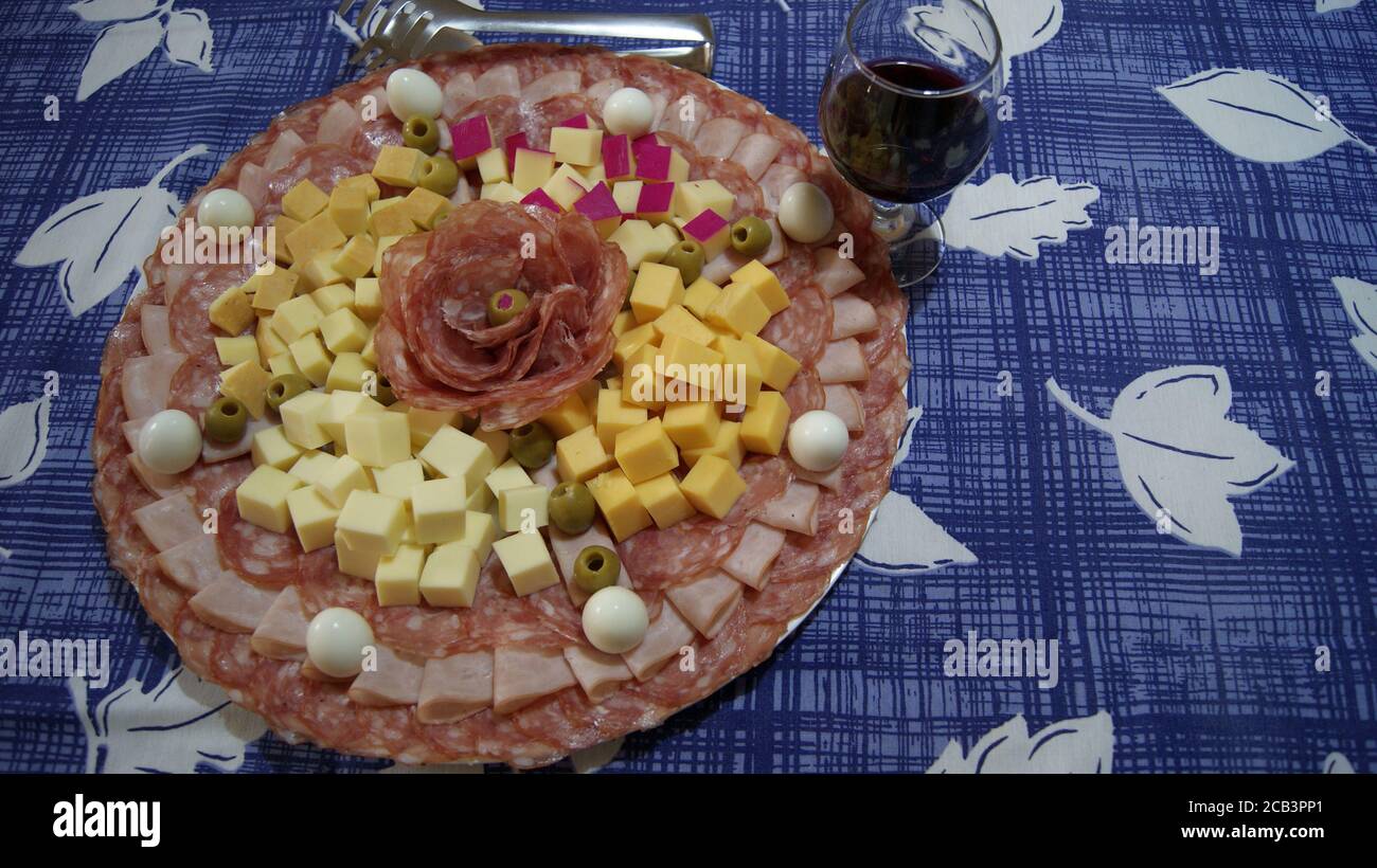 Weißes Gericht mit Salami, Schinken, Wachteleiern und Palmenherz, auf einem Hintergrund mit einer blau-weißen Tischdecke, mit Zange und einem Weinglas Stockfoto