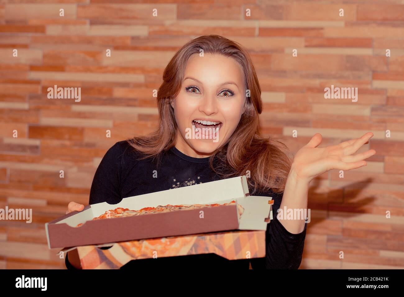 Fröhliche Erwachsene Frau hält Karton mit Pizza und suchen Erstaunt über Kamera auf Holz Wand Hintergrund Stockfoto