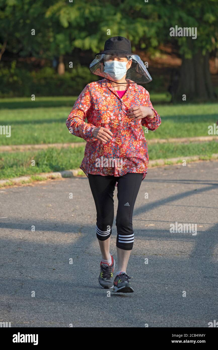 An einem Sommermorgen während der Pandemie, läuft eine Frau trägt eine chirurgische Maske Gesichtsschutz & helle Jacke. In Kissena Park, Flushing, New York City. Stockfoto