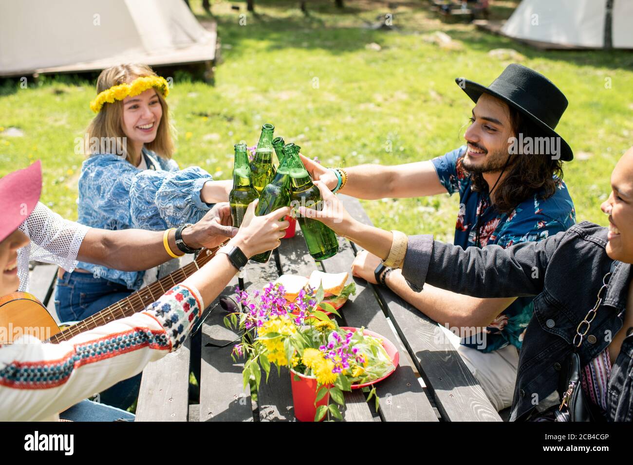 Gruppe von fröhlichen jungen multi-ethnischen Freunden sitzen am Tisch im Freien Und Toasten mit Flaschen auf dem Campingplatz Stockfoto