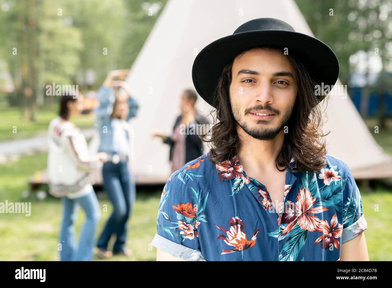Porträt des Inhalts schöner jüdischer Kerl in schwarzem Hut und shirt mit Blumendruck, der mit Freunden auf dem Waldcampingplatz ruht Stockfoto