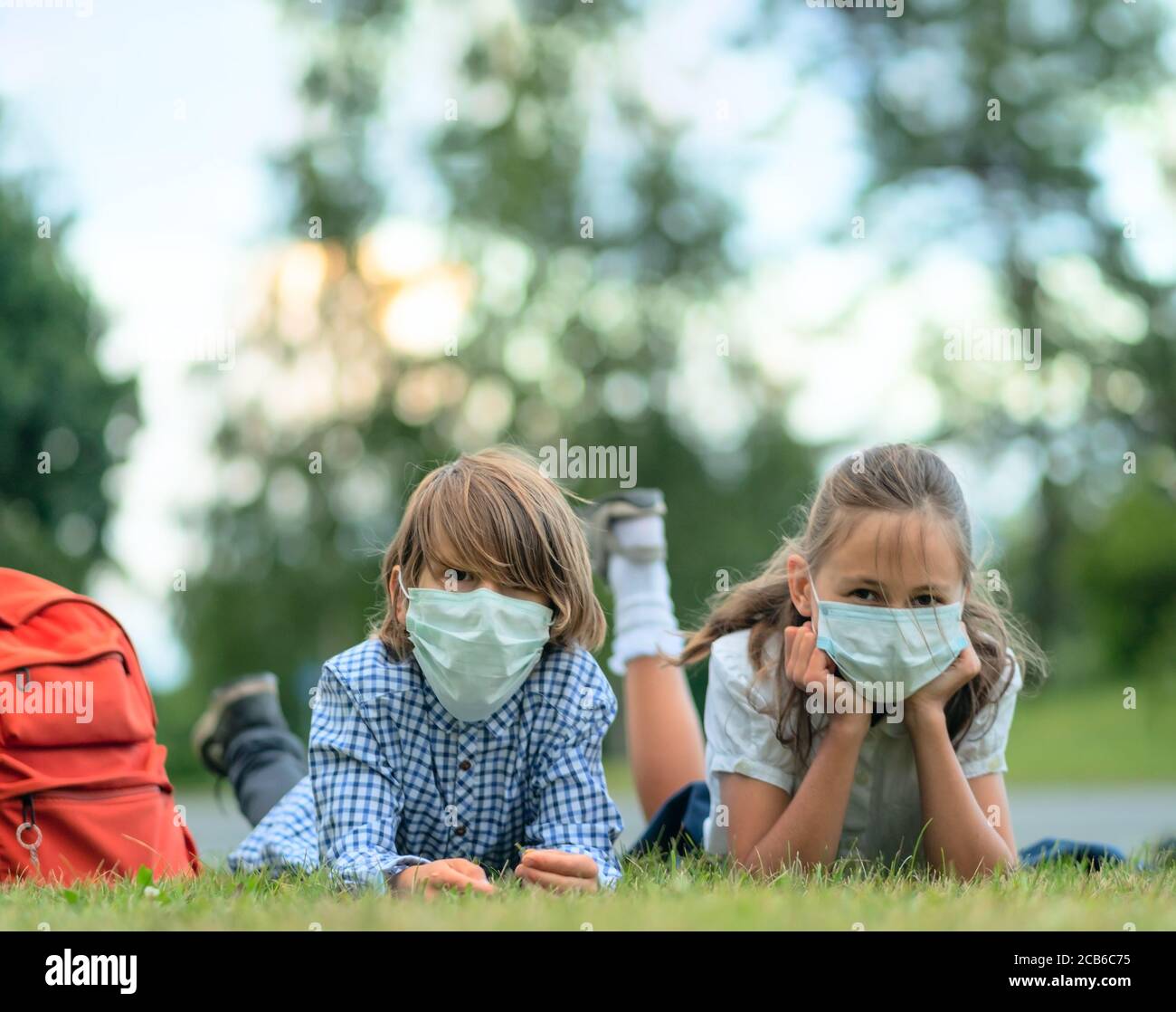 Die Kinder gehen zurück zur Schule. Nette Schüler mit Rucksäcken. Junge und Mädchen in Sicherheitsmasken liegen auf dem Gras. Stockfoto