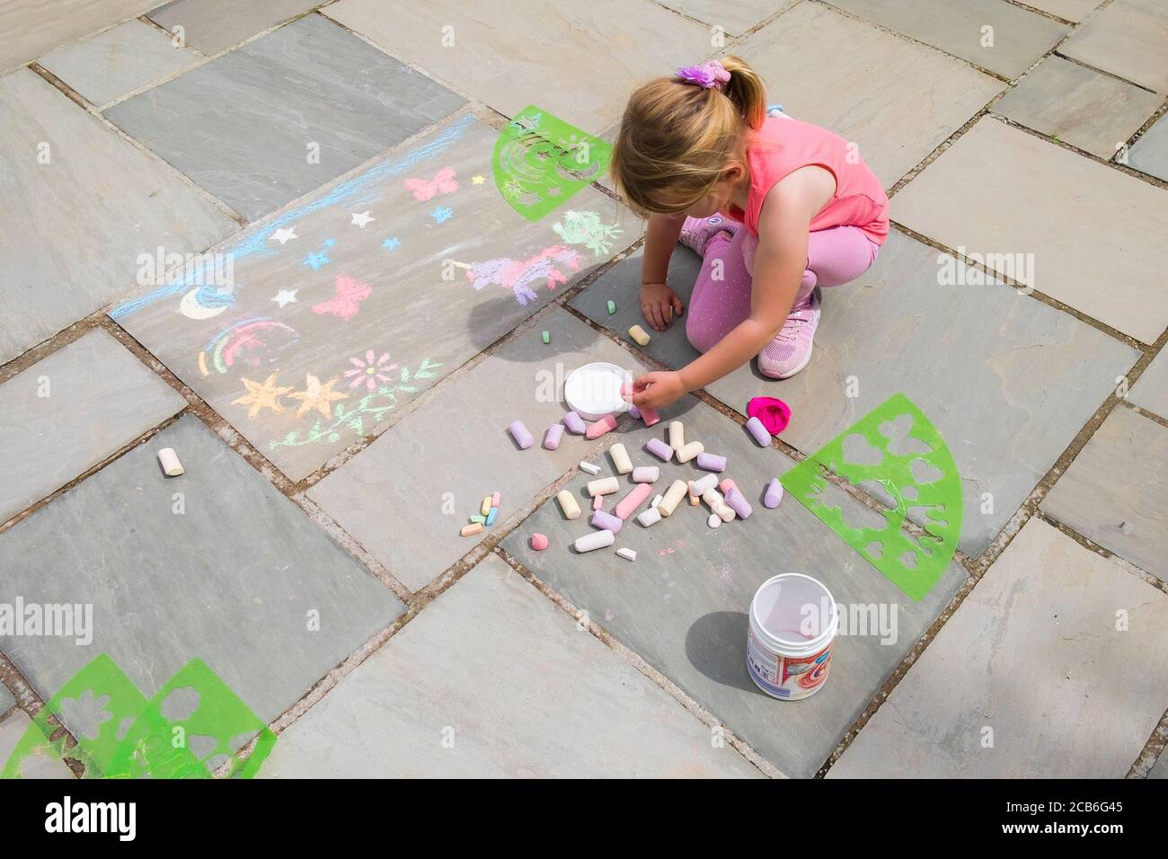 Ein fünfjähriges Mädchen amüsant herslef eifrig mit Plastikschablonen zu Kreide Formen auf einem Plattenboden. Stockfoto