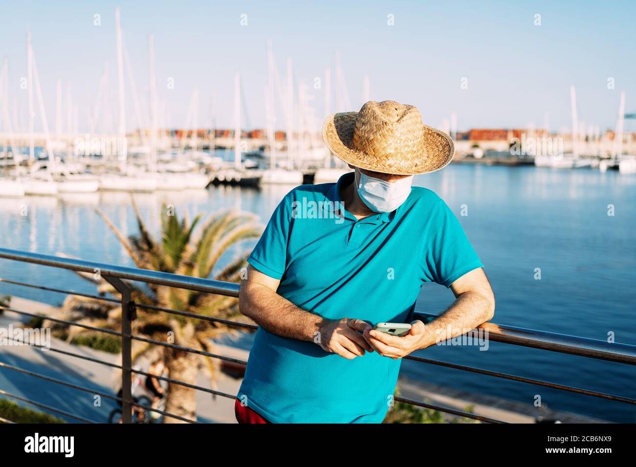 Tourist mit und Gesichtsmaske, die den Inhalt seines Smartphones neben einer Marina bei Sonnenuntergang überprüft. Konzept, persönlicher Schutz in Zeiten von Coronavirus. Stockfoto