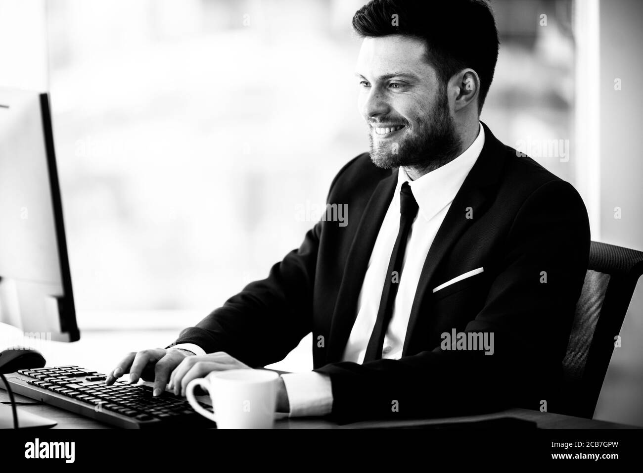 Business Success Konzept. Glücklich lächelnder junger Geschäftsmann sitzt im Büro und arbeitet am Computer. Mann im Anzug drinnen auf Glasfenster Hintergrund Stockfoto